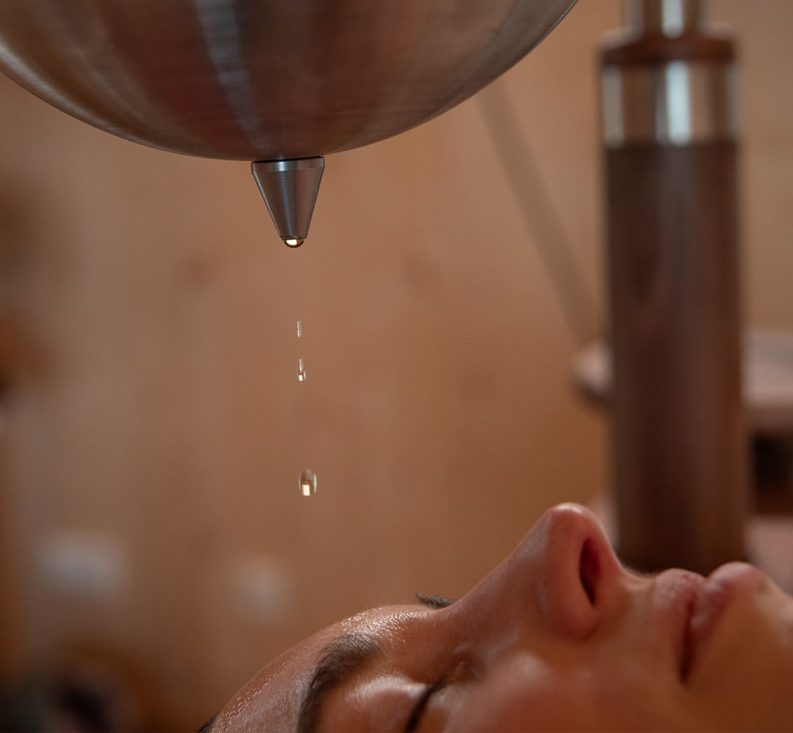 Nahaufnahme des entspannten Gesichts einer liegenden Person mit geschlossenen Augen, auf deren Stirn warme Öltropfen aus einem hängenden Shirodhara-Gefäß fallen. Die Szene, mit einem unschärferen Holzrahmen im Hintergrund, fängt eine tiefe Entspannungsmassage im LIVANDA Zentrum Parin ein.