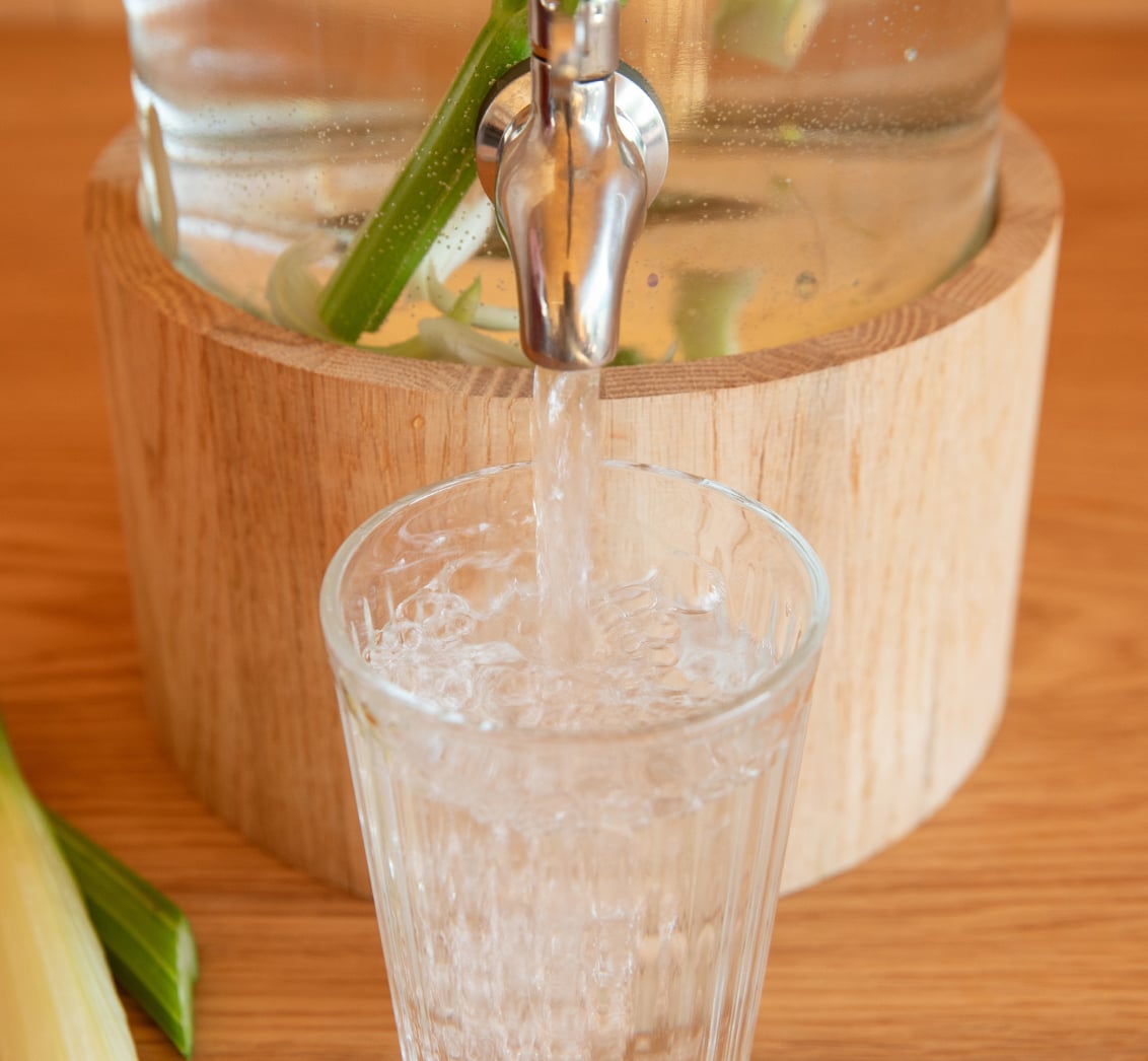 Nahaufnahme eines mit Stangensellerie angereicherten Wasser-Spenders aus Glas, aus dessen verchromtem Zapfhahn Wasser in ein facettiertes Glas fließt. Die Anordnung auf einer Holzoberfläche mit Sellerieblättern im Vordergrund betont das Angebot an frischen, gesunden Erfrischungen für Seminarteilnehmer im LIVANDA Zentrum Parin.