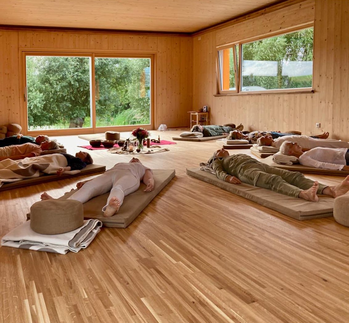 Weitwinkelaufnahme eines hellen, holzverkleideten Seminarraums mit Blick ins Grüne, in dem mehrere Teilnehmer in der Savasana-Haltung auf Matten liegen. Die Komposition, mit einem zentralen Altar aus Klangschalen und Blumen, vermittelt die ruhige Atmosphäre einer Gruppen-Entspannungssitzung im LIVANDA Zentrum Parin