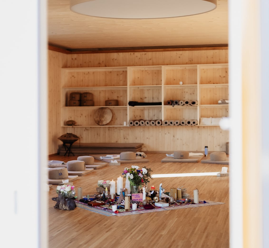 Blick durch eine geöffnete Tür in einen vorbereiteten, holzgetäfelten Seminarraum mit ausgelegten Matten und gestapelten Meditationskissen. Der Fokus auf den zentralen Zeremonienplatz mit Blumen und Kerzen zeigt die Einladung zur Ruhe und Einkehr vor Beginn einer Veranstaltung im LIVANDA Zentrum Parin