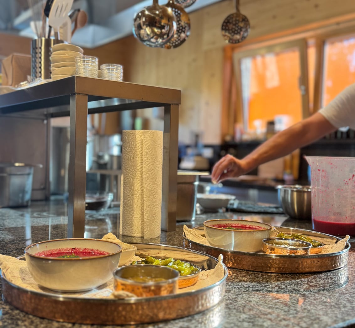 Nahaufnahme von zwei Serviertabletts mit Schüsseln voller Rote-Bete-Suppe, Beilagen und Gewürzen auf einer glänzenden Küchentheke. Die Komposition, mit der unschärferen Küche und dem Koch im Hintergrund, betont das kulinarische Erlebnis und die Verpflegung der Seminarteilnehmer im LIVANDA Zentrum Parin.