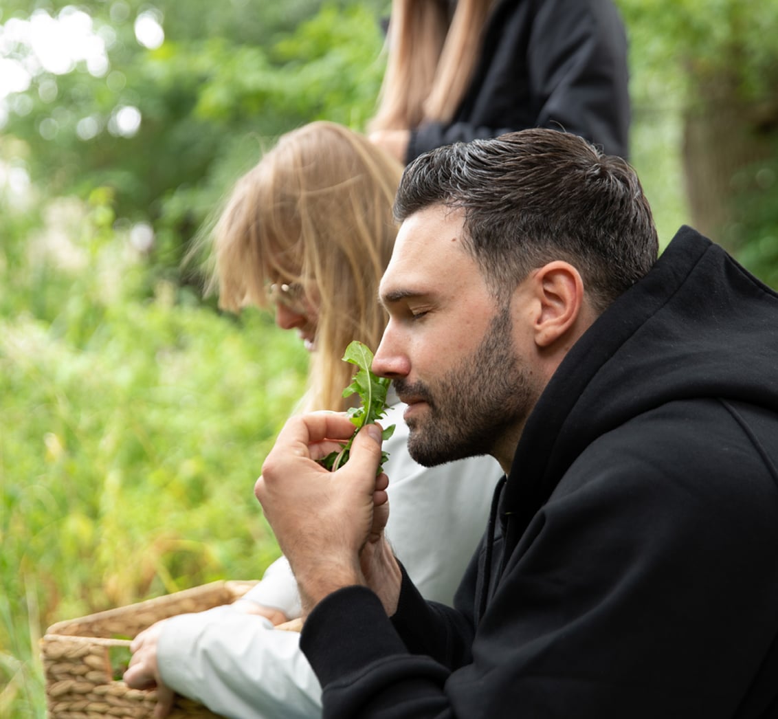 Nahaufnahme eines bärtigen Mannes mit geschlossenen Augen, der konzentriert an frisch gesammelten Löwenzahnblättern riecht. Die Komposition mit einer unschärferen Frau und einem Weidenkorb im Hintergrund fängt die sensorische Erfahrung eines Wildkräuter-Workshops im LIVANDA Zentrum Parin ein.