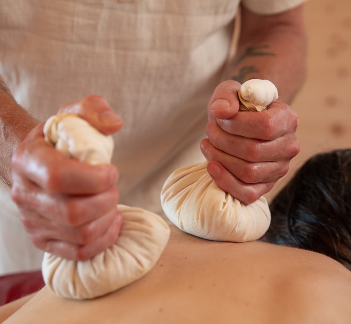 Nahaufnahme der Hände eines Masseurs, der zwei gefüllte Kräuterstempel-Säckchen auf den Rücken einer liegenden Person drückt. Die Komposition mit dem Therapeuten vor einem Holzrahmen fängt eine wärmende Ayurveda-Behandlung im LIVANDA Zentrum Parin ein.