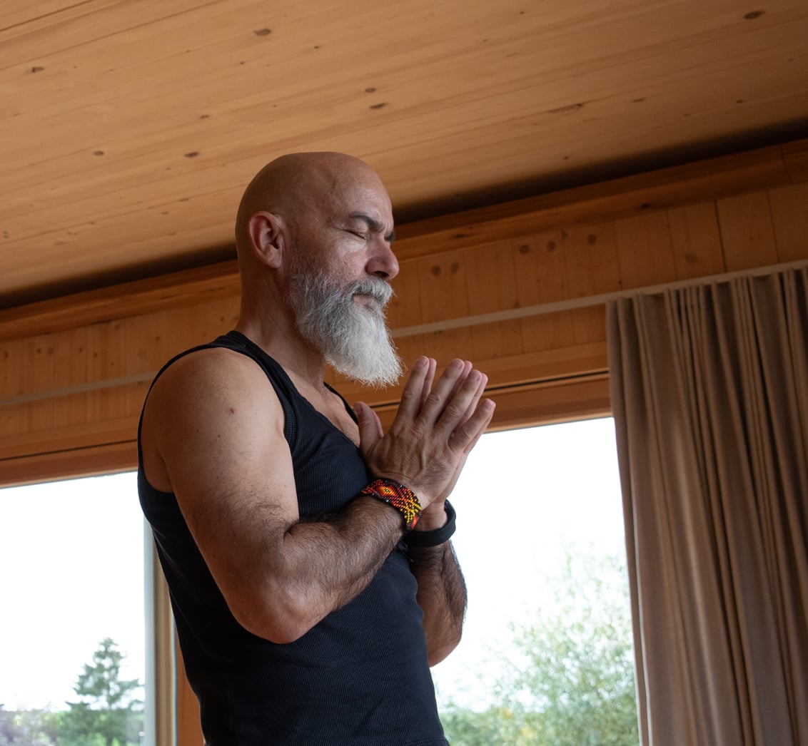 Nahaufnahme eines bärtigen Mannes mit geschlossenen Augen, der in einem holzverkleideten Raum mit gefalteten Händen meditiert. Die Komposition mit dem Mann im Fokus und großen Fenstern im Hintergrund fängt die Stille und Konzentration einer Meditationspraxis im LIVANDA Zentrum Parin ein.