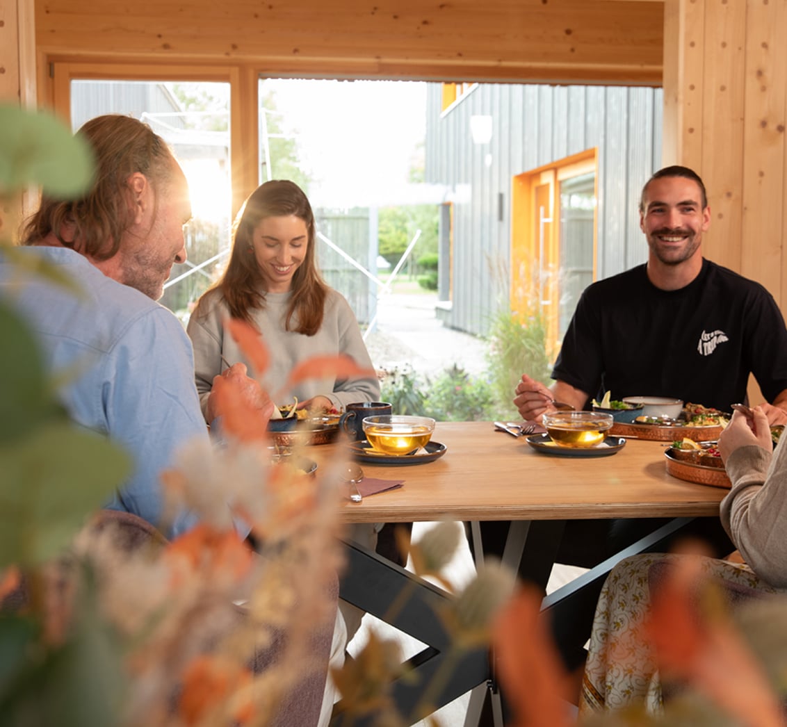 Mittlere Aufnahme einer Gruppe von Seminarteilnehmern, die an einem Holztisch zusammen essen. Die Anordnung mit kupfernen Tellern, Suppenschüsseln und Teegläsern vor einem modernen Gebäude-Hintergrund betont die gemeinschaftliche Atmosphäre und die kulinarische Versorgung im LIVANDA Zentrum Parin.