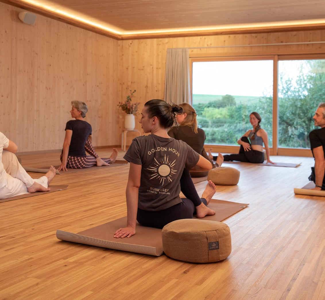 Mittlere Aufnahme einer Yoga-Klasse, in der mehrere Teilnehmer in einem hellen, holzgetäfelten Raum auf Matten sitzen und eine Drehhaltung ausüben. Die Komposition, mit einer Frau mit "GOLDEN HOUR" T-Shirt im Fokus vor großen Fenstern, vermittelt die körperliche Aktivität und die ruhige Atmosphäre eines Yoga-Workshops im LIVANDA Zentrum Parin.