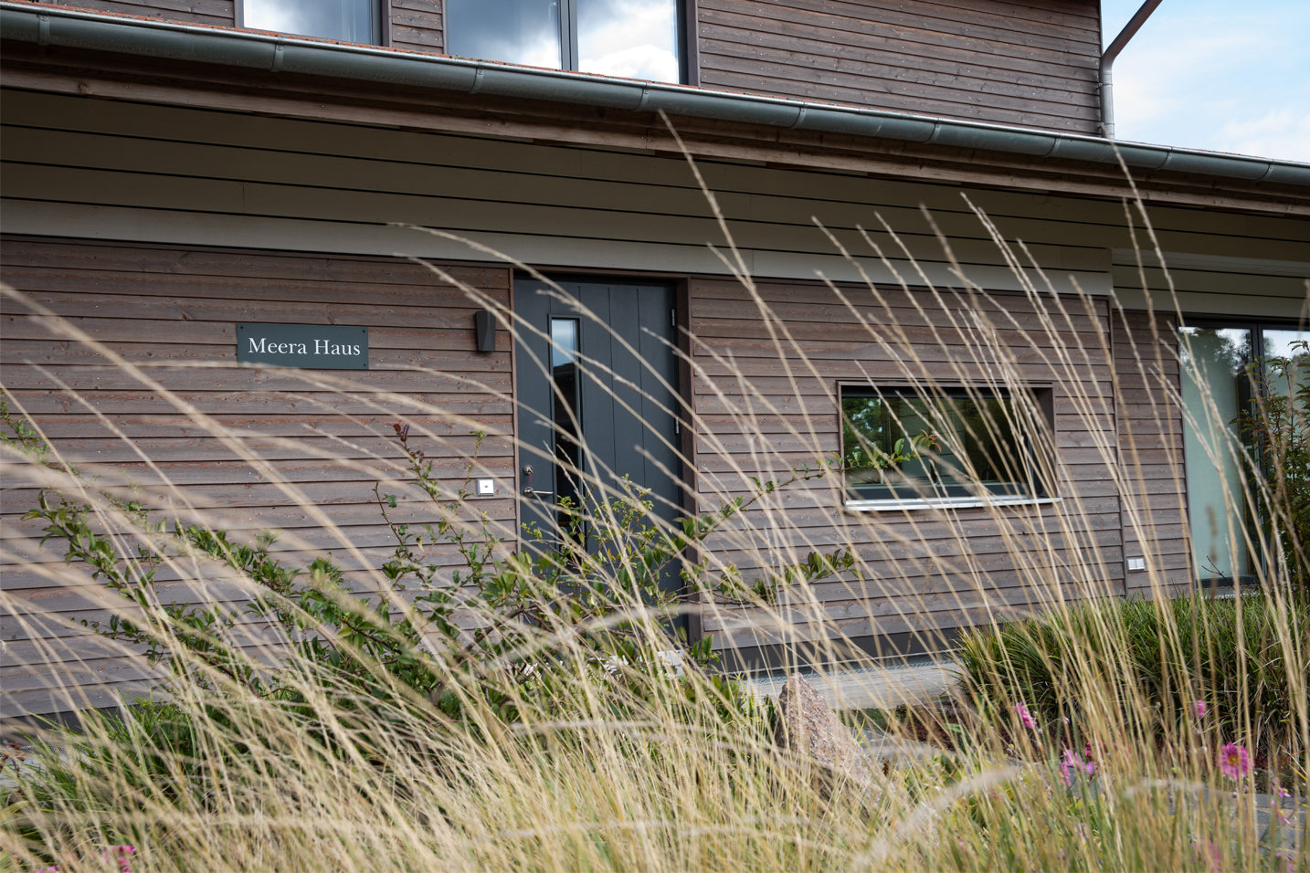 Einladender Eingangsbereich des „Meera Haus“ im LIVANDA Seminarzentrum. Die waagerechte Holzverschalung der Fassade wird von sanft im Wind wehenden Gräsern im Vordergrund eingerahmt, was den natürlichen und friedlichen Charakter des Ortes betont.
