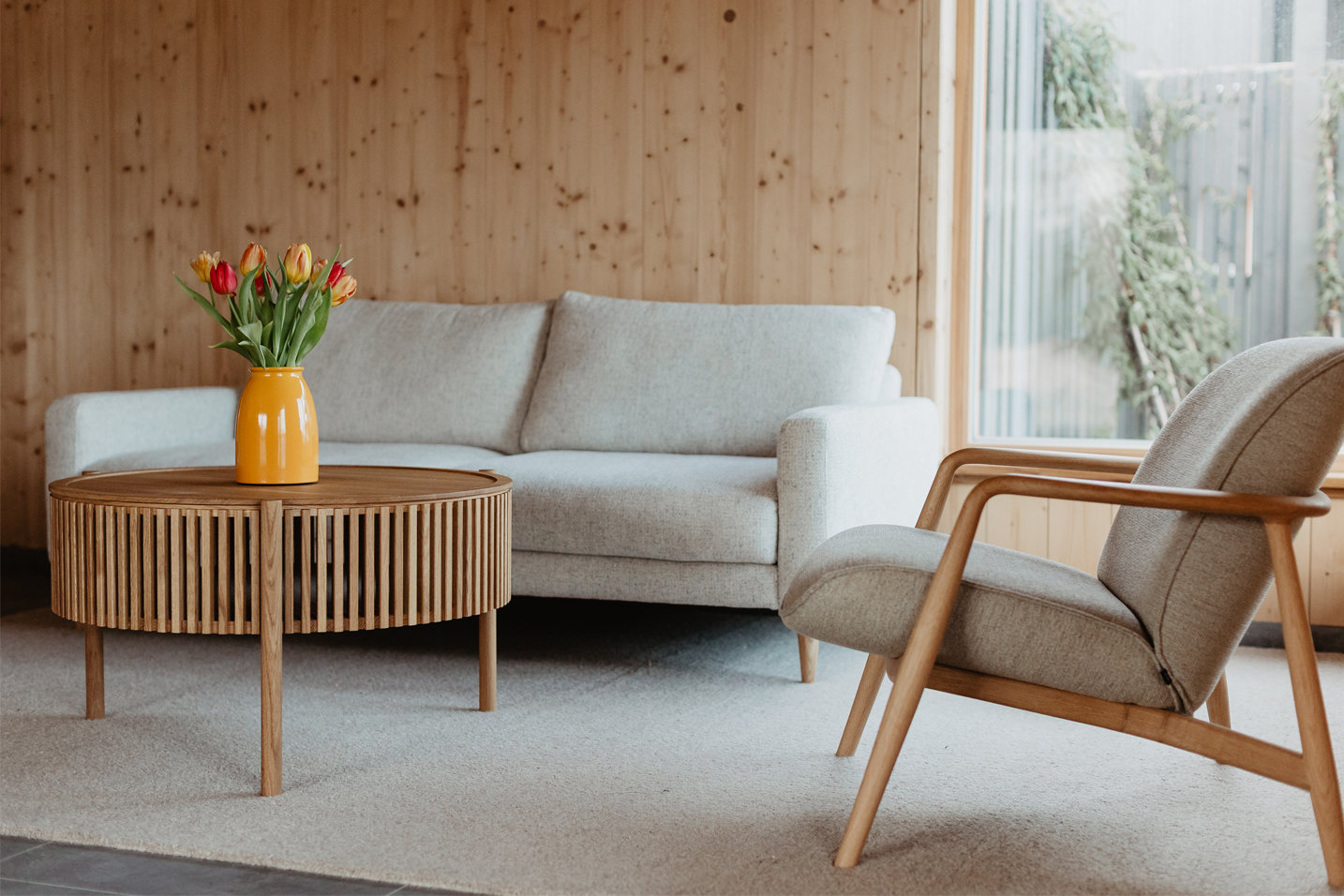 Eine gemütliche Sitzecke im LIVANDA Seminarzentrum, die zum Verweilen einlädt. Ein hellgraues Sofa und ein formschöner Holz-Sessel gruppieren sich um einen runden Echtholztisch. Eine leuchtend gelbe Vase mit bunten Tulpen setzt einen frischen Akzent vor der warmen, hellen Holzvertäfelung und schafft eine Atmosphäre von Geborgenheit.