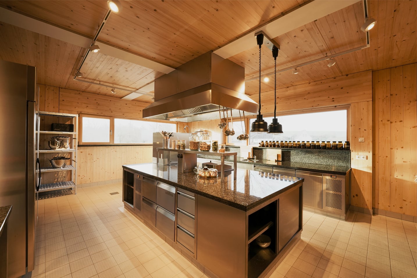 Die moderne und großzügige Gemeinschaftsküche des Zentrums. Eine professionelle Kochinsel aus Edelstahl trifft auf warme Holzwände und hohe Decken. Viel Tageslicht fällt durch die Fensterfront auf die Arbeitsflächen und lädt dazu ein, gemeinsam gesunde Mahlzeiten in einer klaren, inspirierenden Umgebung zuzubereiten.