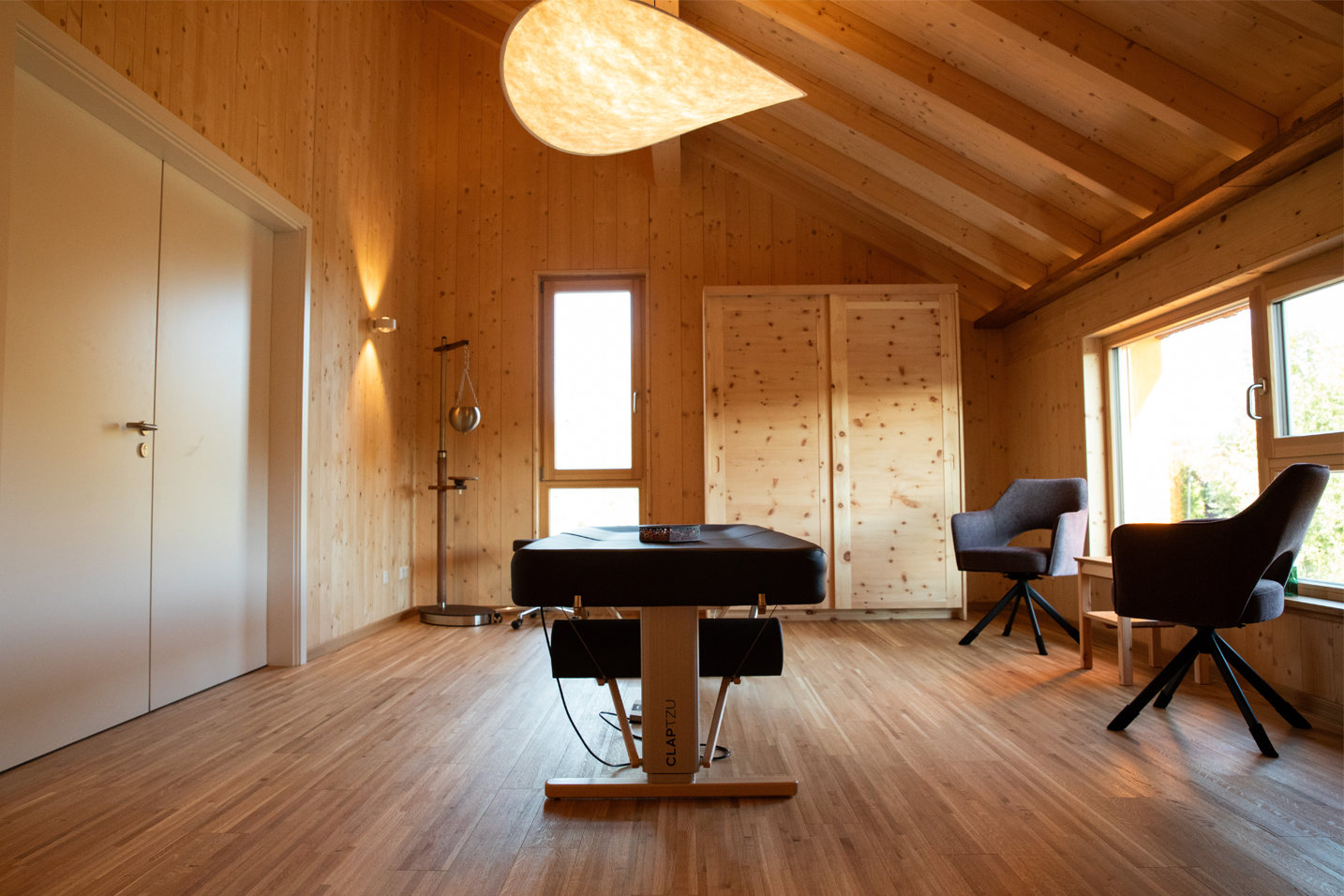 Ein heller, Ruhe ausstrahlender Behandlungsraum mit einer professionellen Massageliege. Die hohen Holzdecken und die sanfte Wandbeleuchtung schaffen einen geschützten Raum für Heilung und Entspannung. Der Blick durch das Fenster ins Grüne unterstreicht die tiefe Verbindung zur Natur.