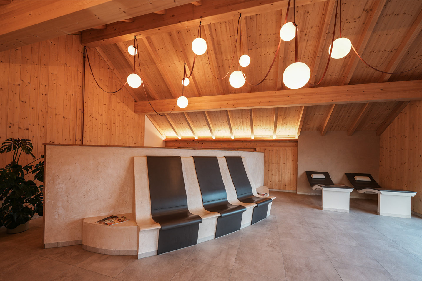 Ein stimmungsvoller Ruheraum im LIVANDA unter einer sanften Dachschräge aus Holz. Moderne, ergonomisch geformte Wärmeliegen laden zur tiefen Entspannung ein. Eine Gruppe von kugelförmigen Pendelleuchten taucht den Raum in ein warmes, weiches Licht, das Geist und Körper zur Ruhe kommen lässt.