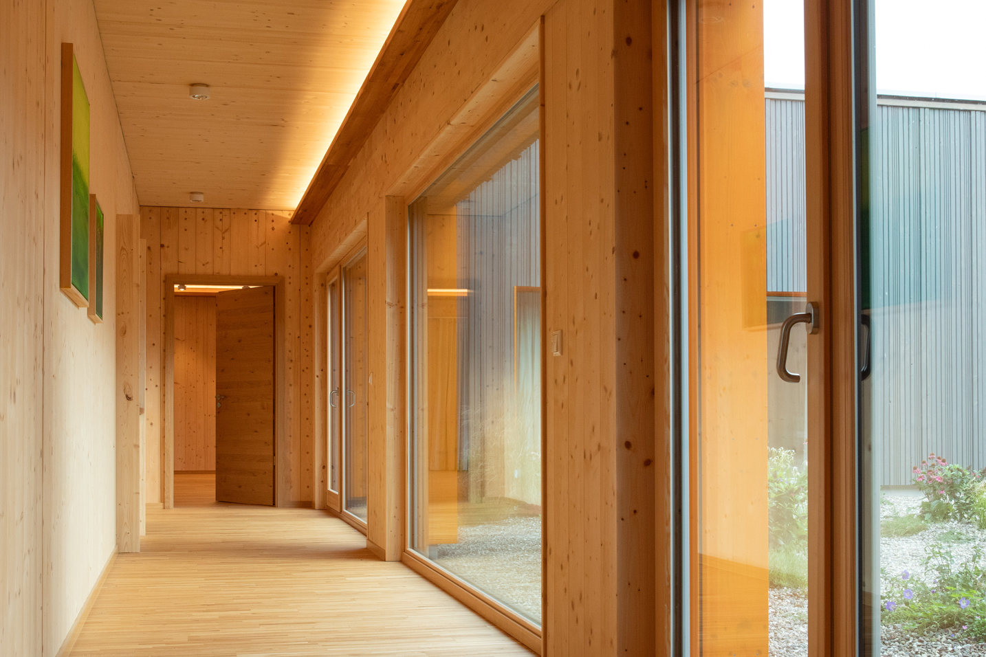 Ein lichtdurchfluteter Flur im LIVANDA, der durch eine raumhohe Glasfront den Blick in den begrünten Innenhof freigibt. Die durchgehende Verwendung von hellem Holz an Boden, Wänden und Decke erzeugt einen fließenden, harmonischen Übergang zwischen Innen und Außen.