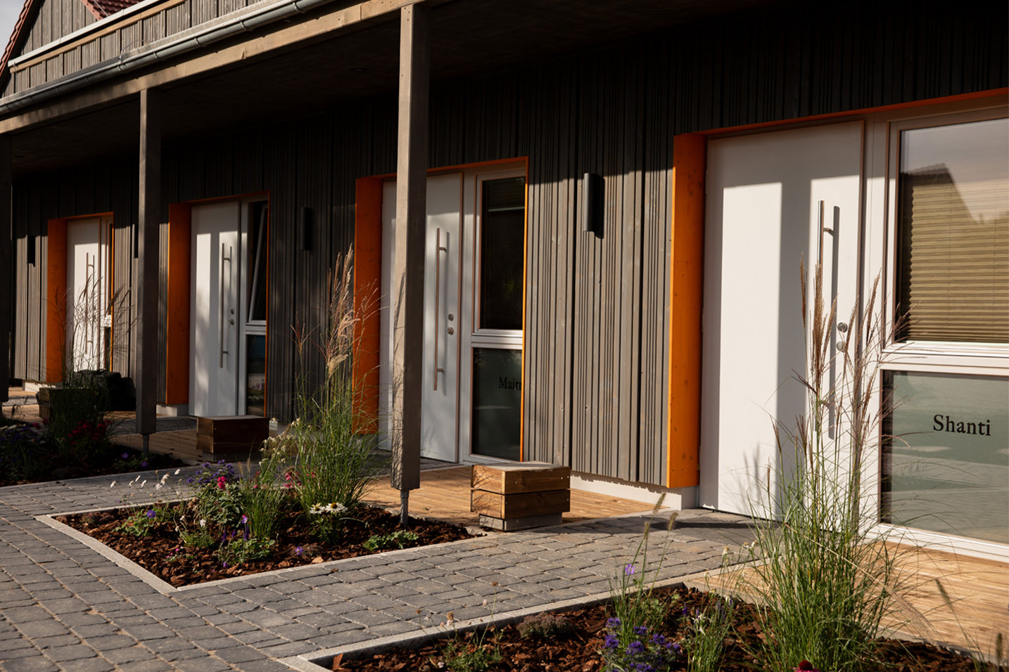 Die Außenansicht der Gästezimmer mit Namen wie „Shanti“ und „Maitri“. Die dunkle Holzfassade, kombiniert mit hellen Türen und kleinen Holzterrassen, wirkt modern und erdend zugleich. Liebevoll angelegte Beete mit Gräsern und Blumen säumen den gepflasterten Weg zu den privaten Rückzugsorten.